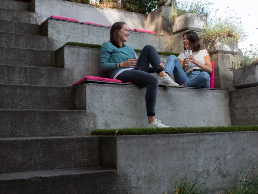 Zwei G&auml;ste sitzen mit Gl&auml;sern auf den Betonstufen im Garten und lachen